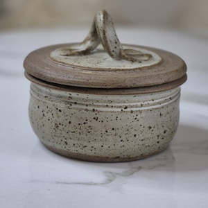 May include: A speckled ceramic jar with a lid. The jar is round with a light beige colour and dark brown speckles. The lid is a darker brown with a loop handle. The jar is on a white surface.
