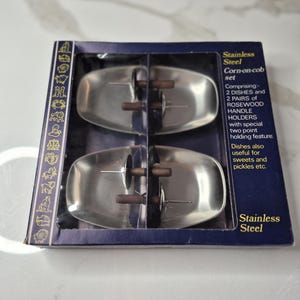 May include: A stainless steel corn-on-cob set, comprising two dishes and two rosewood handle holders. The set is packaged in a dark blue box with text that reads "Stainless Steel Corn-on-cob set". The dishes are also useful for sweets and pickles.