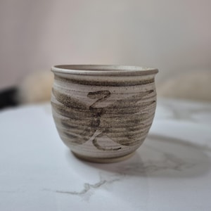 May include: A handmade ceramic cup with a rounded form and a textured, earthy aesthetic. The cup has a light beige base with brown brushstrokes and a dark brown symbol. The rim is a darker shade of brown.