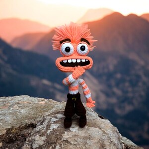 May include: A crocheted toy with orange hair, a wide smile, and brown pants. The toy is standing on a rock with a mountain range in the background.