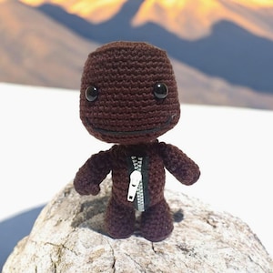 May include: A brown crocheted plush toy with a zipper down the front. The toy has black eyes and a simple smile.