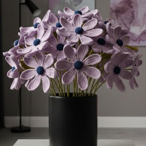 以下が含まれることがあります： 黒い花瓶に入ったかぎ針編みの花のブーケ。ラベンダー色の花は濃い青色の中心と緑色の茎を持っています。円筒形の花瓶は明るい色の表面に置かれています。
