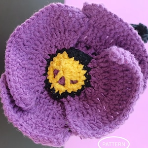 May include: A handmade crochet flower in shades of purple, yellow, and black. The flower has a detailed, textured appearance with a yellow center and black accents. The petals are a deep purple, and the overall design is reminiscent of a poppy or similar bloom.