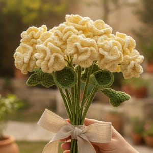Op de afbeelding: Een boeket van gehaakte bloemen in crème- en geeltinten, met groene stelen en bladeren. De bloemen zijn vastgebonden met een beige lint. Een uniek, handgemaakt cadeau.
