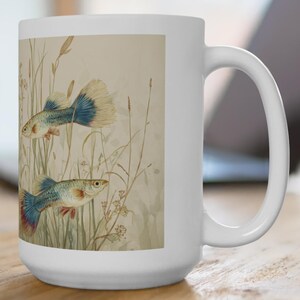 May include: White ceramic mug featuring an illustration of two guppy fish in a naturalistic setting with tall grasses. The fish have blue, red, and yellow details. The mug is on a wooden surface, with a laptop in the background.