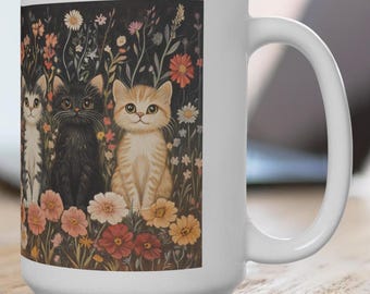 May include: White ceramic mug with a colorful illustration of three cats sitting in a field of flowers. The cats are black, white, and orange and white. The flowers are red, pink, yellow, and purple.
