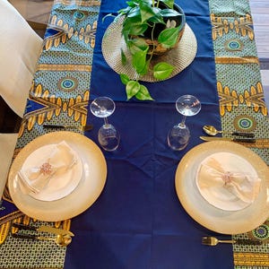 Puede incluir: Un mantel azul con un borde estampado en oro y verde. Dos cubiertos están dispuestos con platos con borde dorado, servilletas blancas y cubiertos de oro. Una planta en maceta se encuentra en el centro de la mesa.