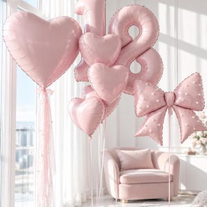 May include: A collection of light pink foil balloons including a large heart, the number 18, three smaller hearts, and a bow shape decorated with white dots. The balloons are attached to thin ribbons and weighted down by small, matching pink balloon weights.