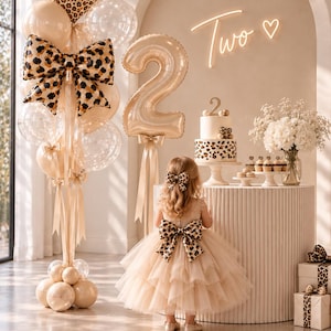 May include: A birthday party display featuring a large balloon bouquet with a leopard print heart and bow, a gold number two balloon, and a tiered cake with leopard print accents. A neon sign reads Two with a heart symbol above a white table.
