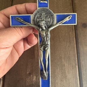 Puede incluir: Un crucifijo de metal plateado con fondo de esmalte azul. El crucifijo presenta una representación de Jesucristo en la cruz, con la inscripción "INRI" sobre su cabeza. La cruz también presenta una medalla circular con la inscripción "CS NOS MD EV-B PAX V-R-S-N-S".
