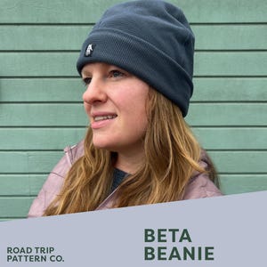 May include: A dark blue beanie with a small white logo is worn by a person. The beanie is made of a textured fabric. The image also includes text that reads "ROAD TRIP PATTERN CO." and "BETA BEANIE". The text also mentions a PDF sewing pattern with 5 sizes.