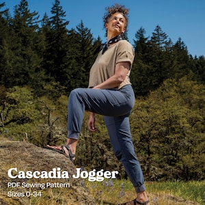 Puede incluir: Una persona con una camiseta beige de manga corta y pantalones deportivos azules está de pie sobre una roca en un bosque. Los pantalones deportivos tienen un tobillo con puños. El texto "Cascadia Jogger" se muestra en blanco, junto con "PDF Sewing Pattern" y "Sizes 0-34".