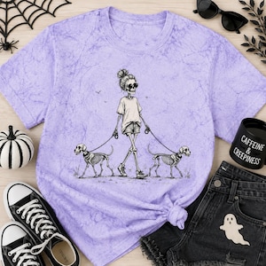 May include: A light purple acid-wash t-shirt featuring a black and white graphic of a skeleton figure wearing sunglasses and shorts, walking two skeleton dogs on leashes. The design is centered on the chest of the short-sleeved crew neck garment.