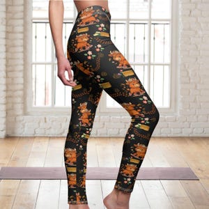 May include: Black leggings featuring a repeating pattern of cartoon Highland cows, flowers, and gold bars. The leggings are form-fitting and made of a stretchy material. The cows are brown and orange with floral crowns.
