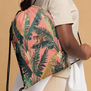 May include: A drawstring backpack with a tropical design. The bag features a pink background with a pattern of green and yellow leaves and a brown monkey. The bag has black drawstrings.