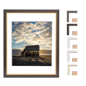 Może przedstawiać: Oprawiony wydruk domu na palach na piaszczystej plaży pod zachmurzonym niebem. Wydruk jest umieszczony w ciemnoszarej ramie ze złotą wewnętrzną obwódką i białym passe-partout. Pokazano kilka próbek narożników ramy.