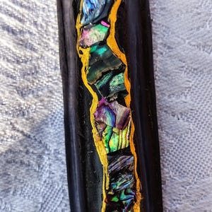 Op de afbeelding: Een zwart, rechthoekig object met een gouden omtrek in de vorm van een zeepaardje. Het zeepaardje is gevuld met iriserende, kleurrijke stukjes abalonschelp. Waarschijnlijk een decoratief item of een kunstwerk.