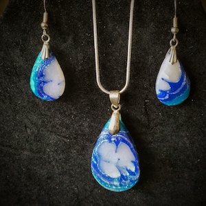 May include: A jewelry set featuring a pendant necklace and matching earrings. The teardrop-shaped pieces have a white and blue marbled design with silver-colored metal accents. The set is displayed on a black surface.