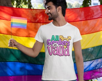 以下が含まれることがあります： 「All Genders are Valid」の文字が緑とピンクでプリントされた白いTシャツ。虹色の旗を手に、より大きな虹色の旗の前に立っています。カラフルなポンポンが付いた青いジーンズを着用しています。