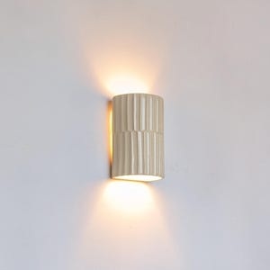 May include: A cylindrical wall sconce with a vertical ribbed texture in a light cream finish. The fixture emits a warm, soft glow from both the top and bottom openings, casting light onto the wall surface above and below the central unit.