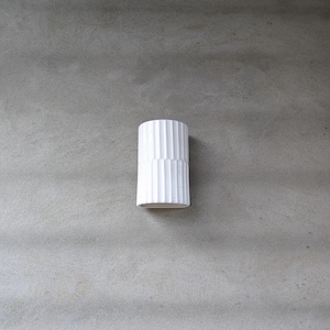 May include: A cylindrical white ceramic vase featuring a vertical ribbed texture throughout its exterior surface. The vase has a consistent diameter from top to bottom with a smooth, matte finish and a clean, open circular rim at the top.