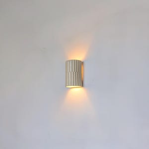 May include: A cylindrical wall sconce featuring a vertical ribbed texture in a matte off-white finish. The fixture emits a warm, golden light from both the top and bottom openings, casting a soft glow against the surrounding wall surface.