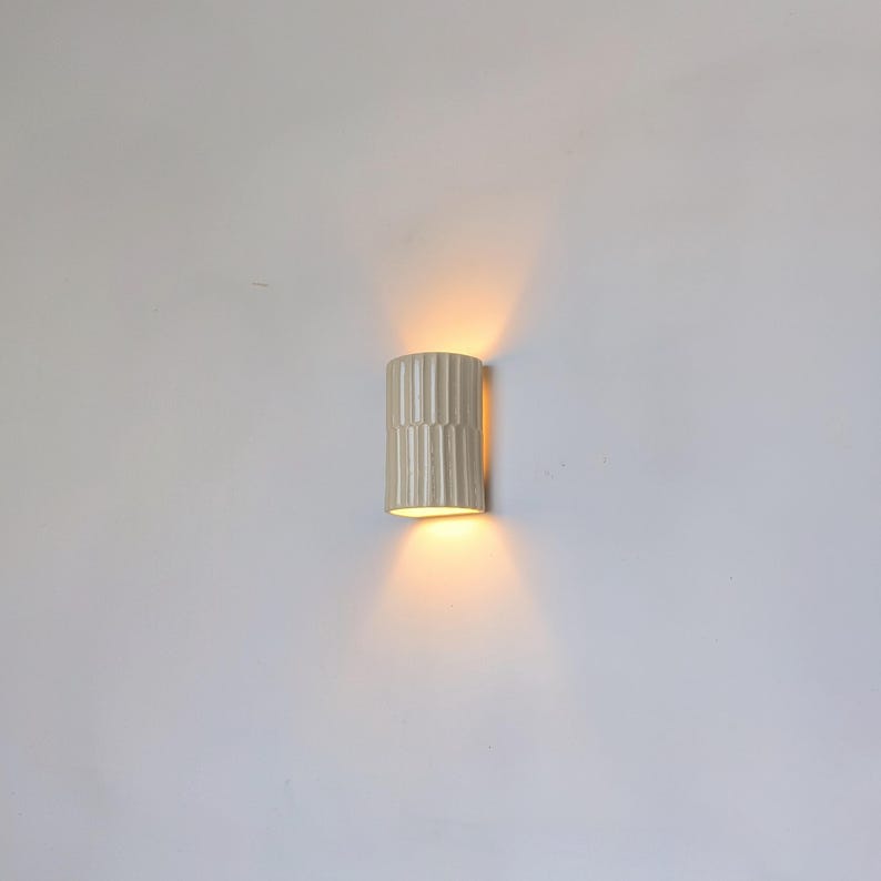 May include: A cylindrical wall sconce featuring a vertical ribbed texture in a matte off-white finish. The fixture emits a warm, golden light from both the top and bottom openings, casting a soft glow against the surrounding wall surface.