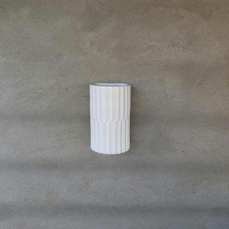 May include: A cylindrical white vase featuring a vertical ribbed texture along its entire exterior surface. The object has a smooth, matte finish and a wide, open top, presenting a clean, uniform appearance with consistent vertical lines throughout.