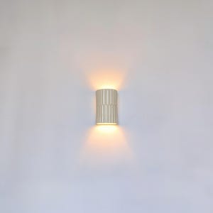 May include: A cylindrical wall sconce with a vertical ribbed texture in a matte off-white finish. The fixture emits a warm, glowing light from both the top and bottom openings, casting soft, symmetrical beams against the surrounding wall surface.