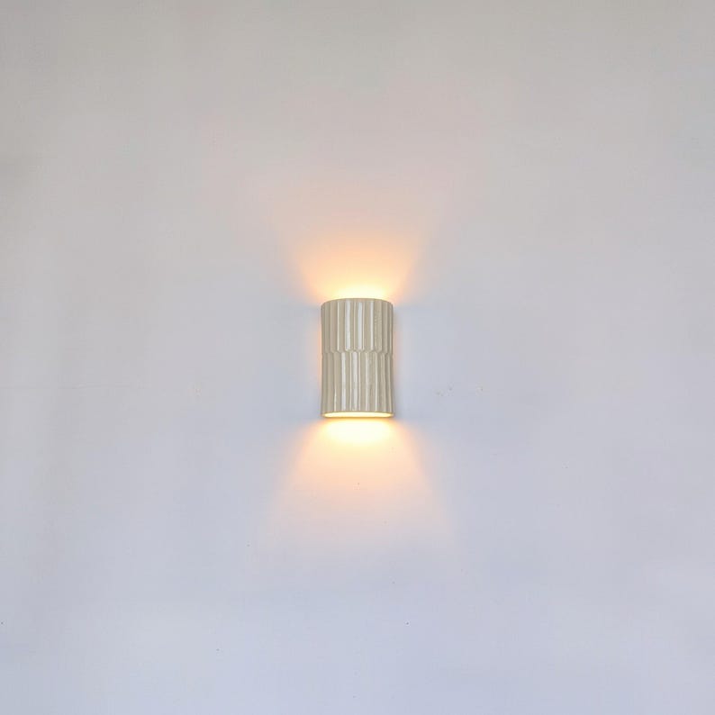 May include: A cylindrical wall sconce with a vertical ribbed texture in a matte off-white finish. The fixture emits a warm, glowing light from both the top and bottom openings, casting soft, symmetrical beams against the surrounding wall surface.