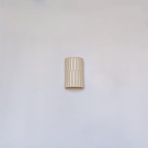 May include: A cylindrical ceramic vase in a light cream color, featuring a vertical ribbed texture that runs along the entire exterior surface. The vase has a smooth, glossy finish and a simple, open top design with a flat base for stability.