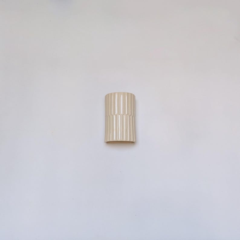 May include: A cylindrical ceramic vase in a light cream color, featuring a vertical ribbed texture that runs along the entire exterior surface. The vase has a smooth, glossy finish and a simple, open top design with a flat base for stability.