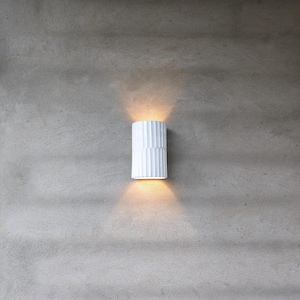 May include: A cylindrical white wall sconce with a vertical ribbed texture. The fixture is mounted on a gray wall and emits a warm, glowing light from both the top and bottom openings, creating two distinct beams of light on the surface.