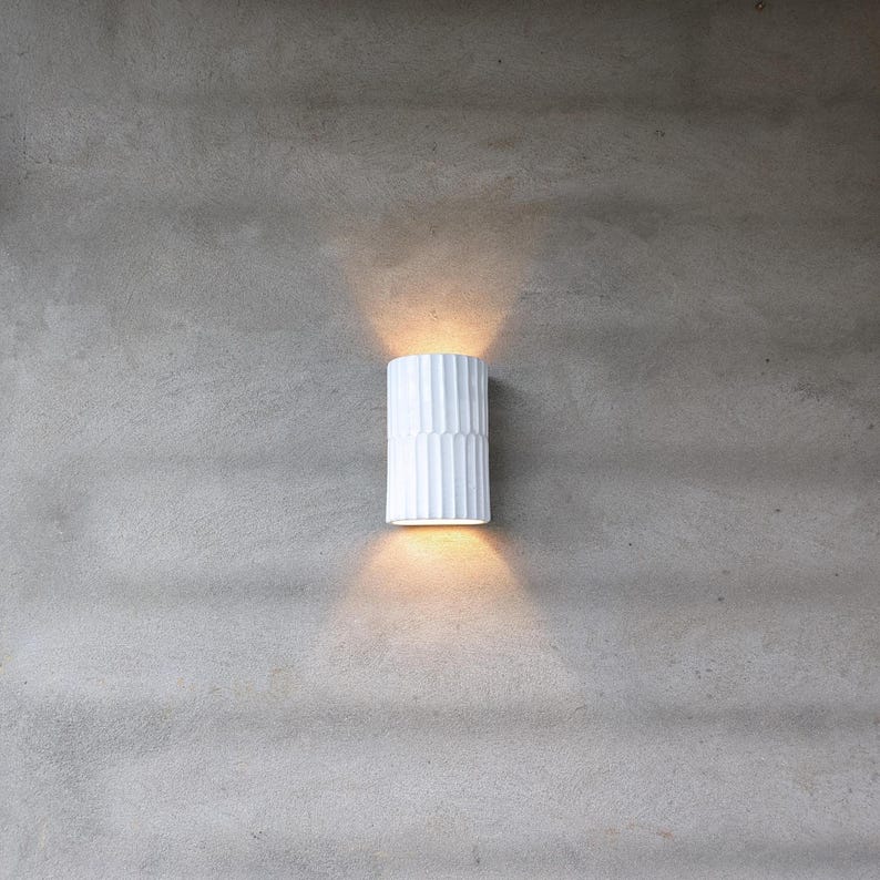May include: A cylindrical white wall sconce with a vertical ribbed texture. The fixture is mounted on a gray wall and emits a warm, glowing light from both the top and bottom openings, creating two distinct beams of light on the surface.