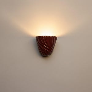 Pode incluir: Um candeeiro de parede vermelho escuro com acabamento brilhante e textura canelada em espiral. A luminária emite um brilho quente voltado para cima que ilumina a parede acima, criando um efeito de luz suave com uma forma simples.