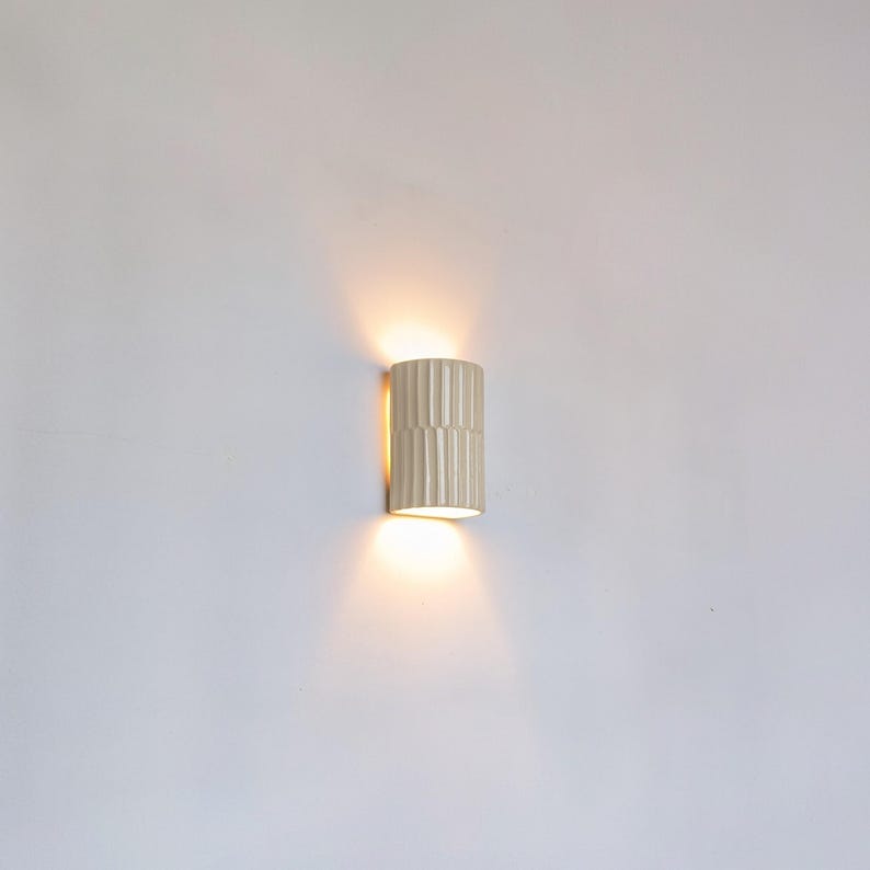 May include: A cylindrical wall sconce with a vertical ribbed texture in a light cream finish. The fixture emits a warm, soft glow from both the top and bottom openings, casting light onto the wall surface above and below the central unit.