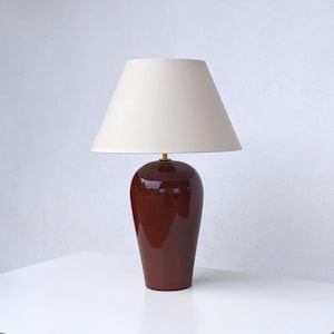 Peut inclure: Une lampe de table avec un pied en céramique bordeaux et un abat-jour crème. Le pied a une finition brillante et une forme arrondie. L'abat-jour est conique. La lampe est sur une surface blanche.