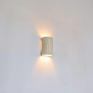 May include: A cylindrical wall sconce with a vertical ribbed texture and a glossy off-white finish. The fixture emits a warm, soft glow from both the top and bottom openings, casting light onto the wall surface above and below the unit.