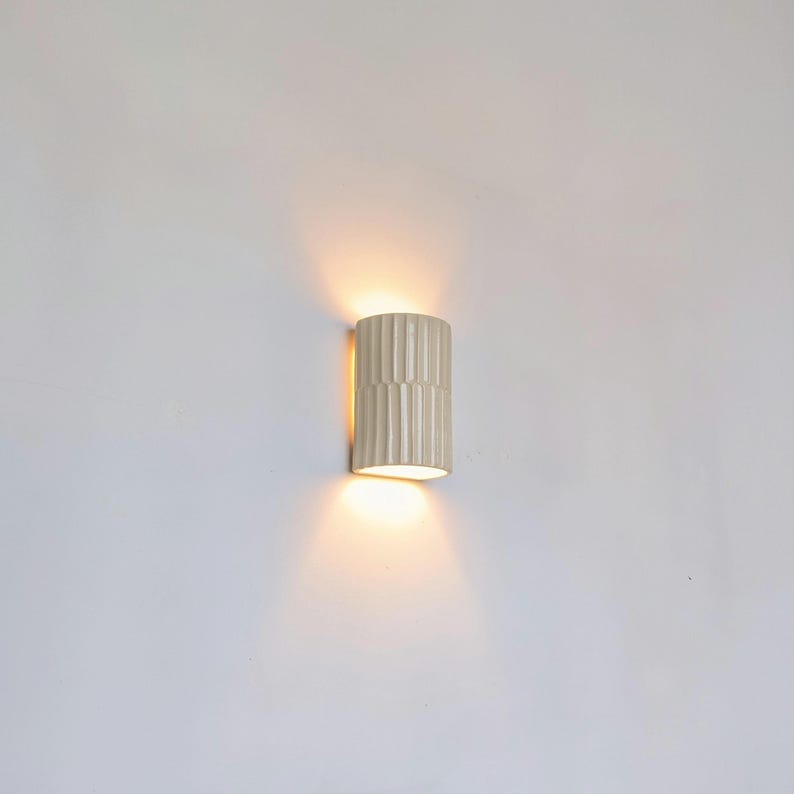 May include: A cylindrical wall sconce with a vertical ribbed texture and a glossy off-white finish. The fixture emits a warm, soft glow from both the top and bottom openings, casting light onto the wall surface above and below the unit.
