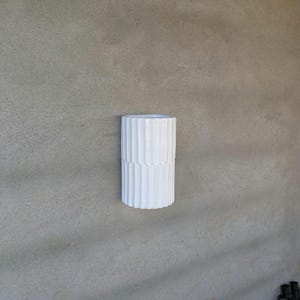 May include: A white ceramic wall planter with a cylindrical shape and a vertical ribbed texture. The planter features a flat top opening and a smooth, glossy finish, designed to be mounted flush against a vertical surface for display purposes.