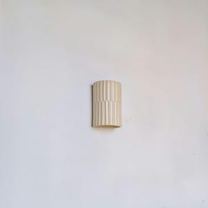 May include: A cylindrical ceramic wall sconce in a light cream color, featuring a vertical fluted texture along its entire exterior surface. The fixture has a smooth, glossy finish and a semi-circular shape designed to mount flush against a wall.
