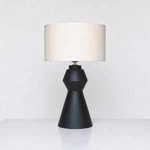 Puede incluir: Una lámpara de mesa con una base negra mate de silueta geométrica y escalonada, junto a una pantalla cilíndrica de tela color blanco roto. La lámpara presenta una pequeña pieza de conexión color latón entre la base y la pantalla.