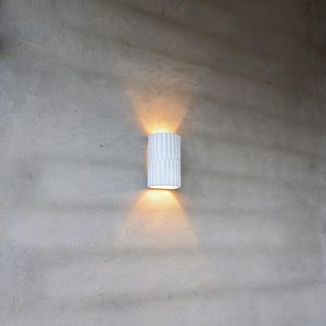 May include: A cylindrical wall sconce with a white, vertically ribbed exterior surface. The fixture emits a warm, golden light from both its top and bottom openings, casting two distinct, symmetrical beams of light onto the surrounding wall surface.