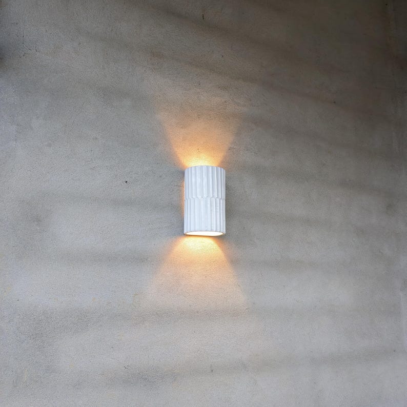 May include: A cylindrical wall sconce with a white, vertically ribbed exterior surface. The fixture emits a warm, golden light from both its top and bottom openings, casting two distinct, symmetrical beams of light onto the surrounding wall surface.