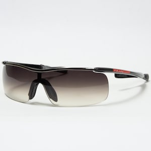 May include: Black and gray Prada sunglasses with a gradient lens. The sunglasses have a sporty design with a black frame and a red accent on the temple. The lenses transition from dark to light.