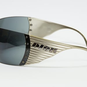May include: Silver-toned sunglasses with a wraparound design. The lenses are a dark gray, and the frames feature a ribbed texture with small rivets. The temples curve gracefully, and the brand name is visible on the side.