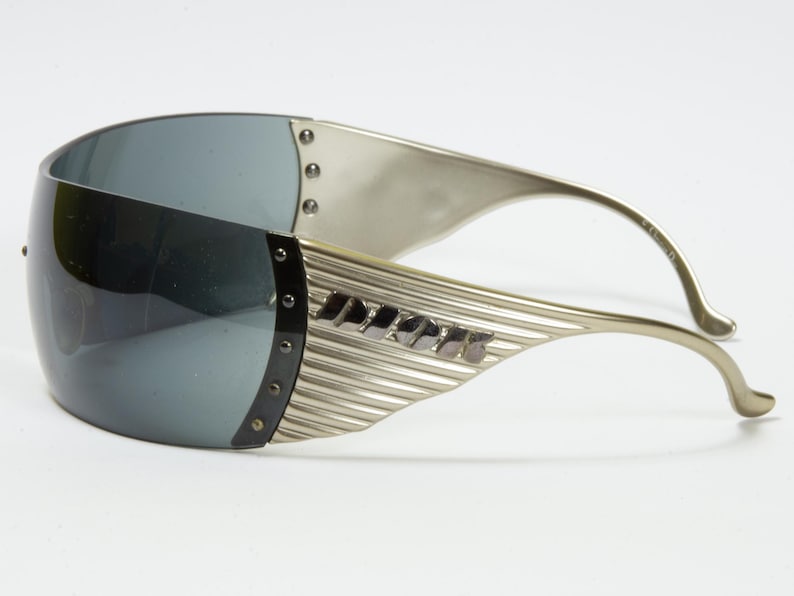 May include: Silver-toned sunglasses with a wraparound design. The lenses are a dark gray, and the frames feature a ribbed texture with small rivets. The temples curve gracefully, and the brand name is visible on the side.