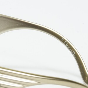 May include: Close-up of a pair of gold-toned eyeglasses. The temple arm is curved and features the text "&copy; Christian Dior" in a delicate font. The glasses have a sleek, modern design.