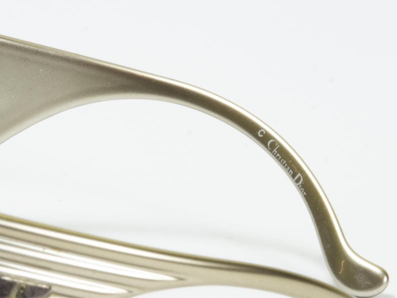 May include: Close-up of a pair of gold-toned eyeglasses. The temple arm is curved and features the text "&copy; Christian Dior" in a delicate font. The glasses have a sleek, modern design.