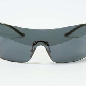 May include: A pair of shield sunglasses with a dark gray lens and a curved, wraparound design. The frame is a dark color, and small decorative studs line the outer edges. The sunglasses have a bridge and arms.
